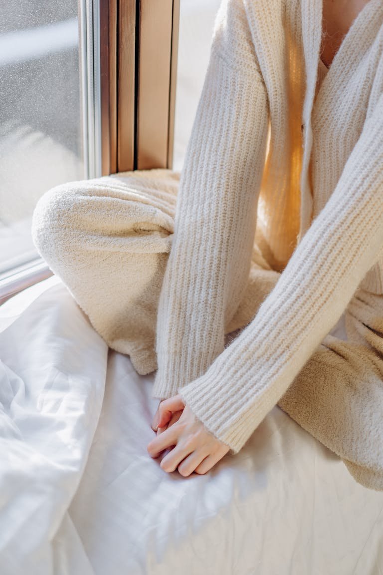 Woman in a cozy sweater sits by a sunlit window, exuding warmth and relaxation.